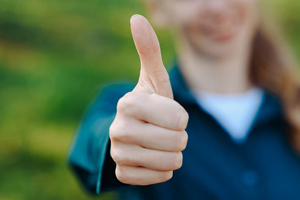 young beautiful redhead caucasian woman shows thumb up gesture with fingers, green shirt, city park in sunny summer day, smiling happily