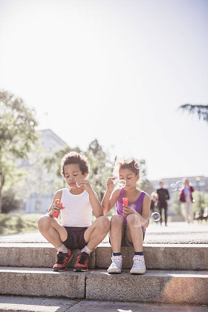 Two children, a boy and a girl, are joyfully sitting on outdoor steps, blowing bubbles and enjoying a sunny day.