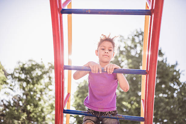 A young child wearing a purple shirt climbs colorful playground equipment on a sunny day, with trees in the background.