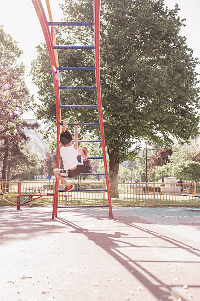 A child is enjoying a sunny day at the park, climbing on a red jungle gym ladder. The light filters through the trees, casting shadows on the ground.