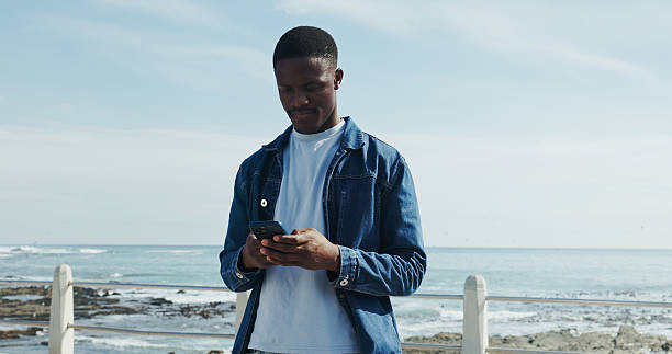 istock Scroll, message and man at promenade with phone, communication or typing on mobile app. Concentration, digital and African male at beach with tech, connection and text update on social media. 2238434709