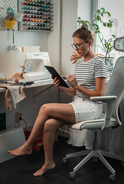 istock Woman tailor relaxes in cozy home workspace while planning on tablet next sewing project. Warm atmosphere and ergonomic setup support her focus on handmade clothing production. Wall board, side view. 2238450568