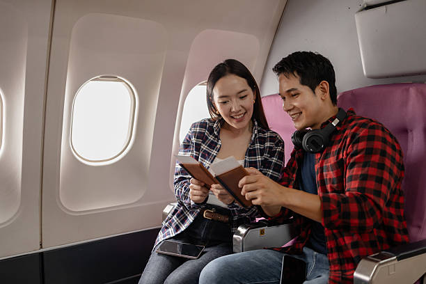 istock Asian couple sitting together on an airplane, smiling while looking at documents. They appear happy and excited about traveling, dressed casually with a bright airplane cabin in the background. 2238451205