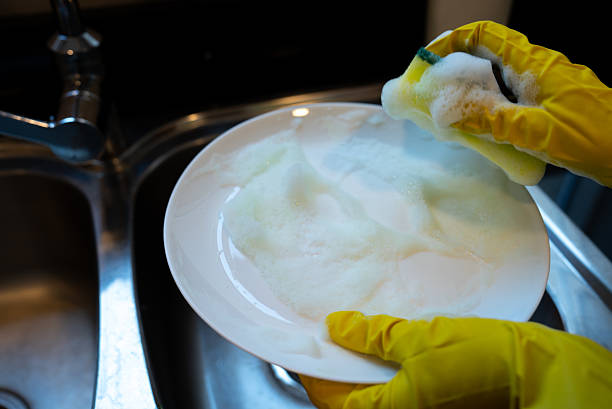 High-contrast close shot of a plate full of white soap suds being cleaned by a gloved hand. Fresh, bubbly domestic hygiene imagery.