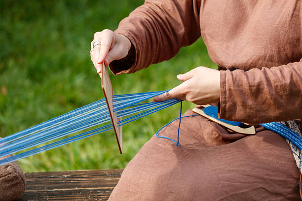 Man is sitting on a bench and is weaving a blue and white string. He is holding a wooden tool in his hand
