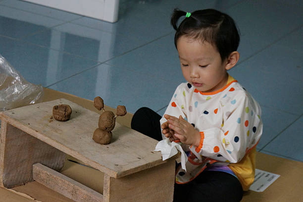 Thai child focused on playing with clay, engaged in the process of creation. The child is seated at a wooden table, molding the clay with focus and care