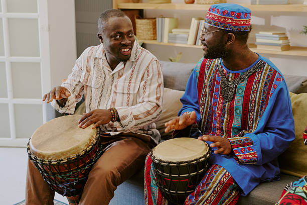 Two middle aged Black men sitting on sofa playing traditional drums and smiling at each other during Kwanzaa celebration, wearing patterned clothing, engaging in cultural activity
