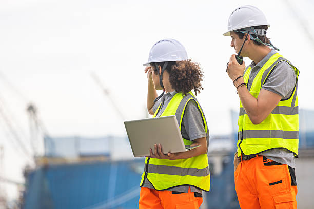 istock Construction Workers Reviewing Site Plans with Laptop and Radio, Two Industrial Workers in Safety Gear on Port Site, Teamwork and Communication Engineers with Laptop and Walkie-Talkie on Site 2238500422