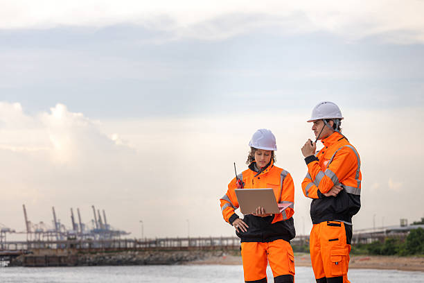 istock Engineers or Maritime Workers Reviewing Data with a Laptop and Radio at a Port, Two Professionals in High-Visibility Safety Gear Outdoors, Technical Workers Overseeing Operations by the Water 2238502906