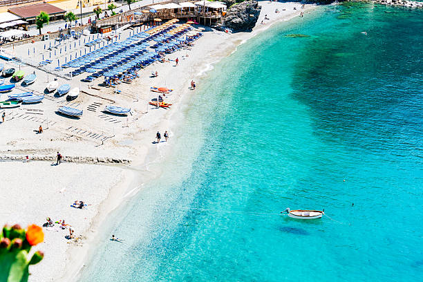 Monterosso al Mare beach with distinctive large rocks on the shoreline, white sandy coast where people relax under umbrellas, and clear turquoise water with surrounding hills in the distance.