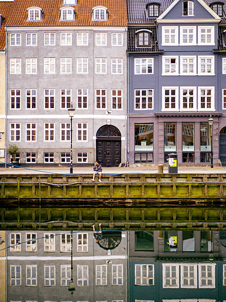 Copenhagen, Denmark – August 2, 2025: Traditional colorful facades of buildings in Nyhavn, with a person sitting on the canal edge and the structures clearly reflected in the water.