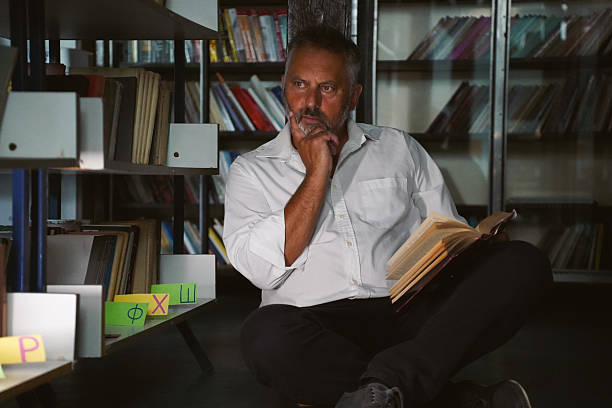 Older man in a white shirt browsing shelves, picking and reading books in the library. Theme of education, research, and love for reading in a quiet space with books helve