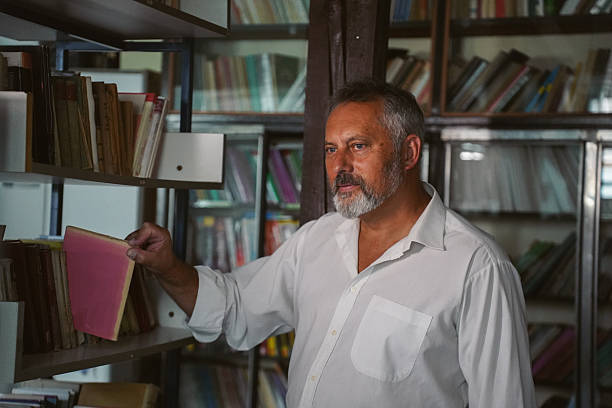 Older man in a white shirt browsing shelves, picking and reading books in the library. Theme of education, research, and love for reading in a quiet space with books helve