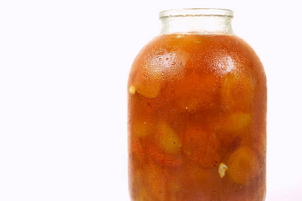 A jar filled with vibrant orange marmalade rests on a kitchen counter, glistening in the sunlight. The thick texture and fruit pieces suggest a homemade delight ready to be enjoyed.
