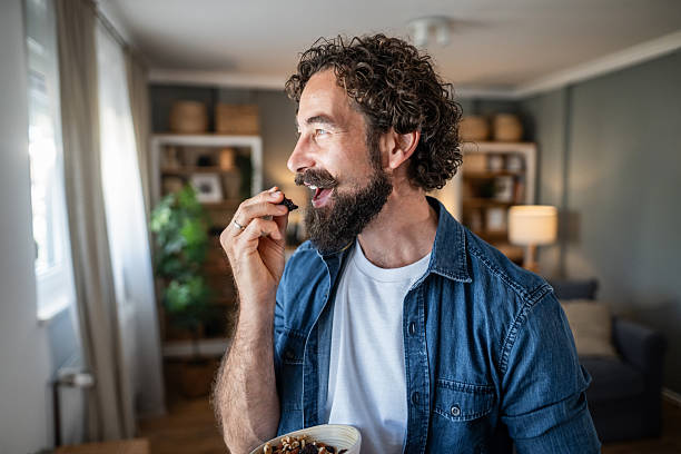 Bearded man with curly hair in denim shirt enjoys a mix of dried fruits and nuts while looking out a window in his modern home, content and practicing mindful healthy eating