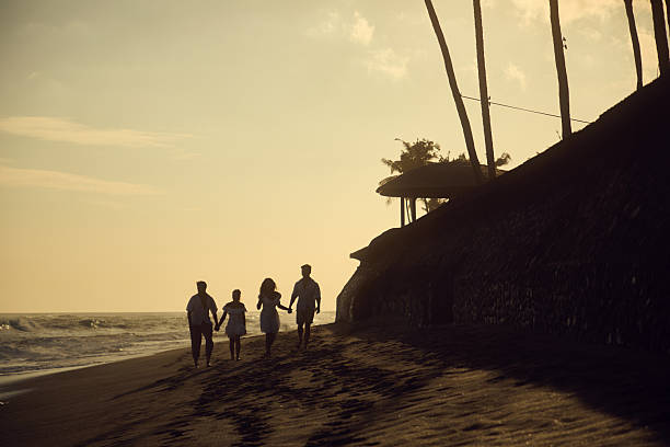 A group of four silhouetted against the vibrant hues of a beach sunset, enjoying the warmth and peacefulness of the evening. Palms and a distant hut add to the tranquility.