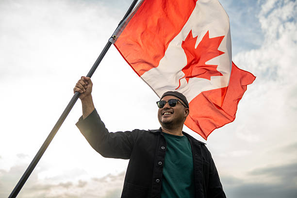 Portrait of cheerful young man looking forward while standing proudly with holding canada flag over his head against wind and blue sky Man holding the canada flag celebration
