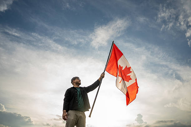 istock Portrait of cheerful young man looking forward while standing proudly with holding canada flag over his head against wind and blue sky Man holding the canada flag celebration 2238535403