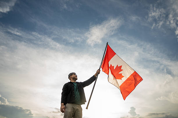 istock Portrait of cheerful young man looking forward while standing proudly with holding canada flag over his head against wind and blue sky Man holding the canada flag celebration 2238535410