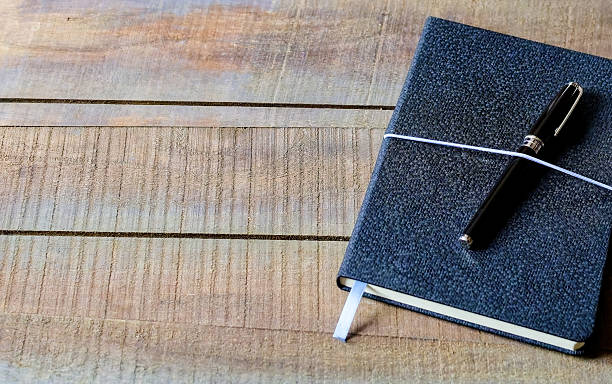 On a light brown wooden table, a dark blue diary sits with a black pen across it, held by a white string for a therapy session. A white bookmark sticks out of the diary. Copy space for text, high angle view - Mental health, expressive writing