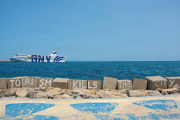 Palermo, Italy - May 18 2025. A protest message against the impact of mass tourism, Tourism Kills the City, written on the breakwater at Palermo waterfront. In the background, a large cruise ship sails to port, a juxtaposition representing one of the things being protested against, highlighting the tension between overtourism and local needs. Located at the Esplanade or Lungomare waterfront, in Foro Italico in the Kalsa district