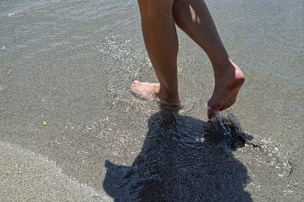A close-up view captures a pair of bare human feet standing in shallow, clear ocean water on a wet sandy beach. The feet are positioned ankle-deep, with one foot slightly lifted as gentle waves lap around them, creating ripples in the sand. A prominent shadow of the person stretches across the foreground on the damp sand. The scene conveys a sense of calm and immersion in nature, with natural sunlight casting the shadow during daytime. No full figures are visible, emphasizing the textures of skin, water, and sand. This image suits commercial applications such as travel brochures promoting coastal destinations, lifestyle content on relaxation and mindfulness at the seaside, photography tutorials illustrating shadow play in outdoor shoots, summer advertising for beachwear or vacations, and editorial pieces on personal exploration or nature photography. The outdoor setting suggests a temperate coastal location, ideal for themes of leisure and creative pursuits.