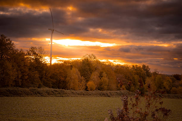 In a sky full of dramatic clouds and glowing orange light. The autumn-colored trees below reflect the warm shades of the Golden Hour. The picture contrasts modern technology with the enormous power of nature.