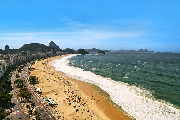 The shadows of a couple standing close together are cast on the wet sand of a beach in Urca, Rio de Janeiro. The sea reflects the strong midday light, with white waves rolling toward the shore.