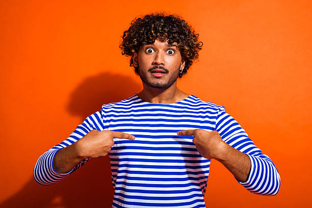 istock Young man in striped shirt expressing surprise while pointing at himself against bright orange background 2238566437