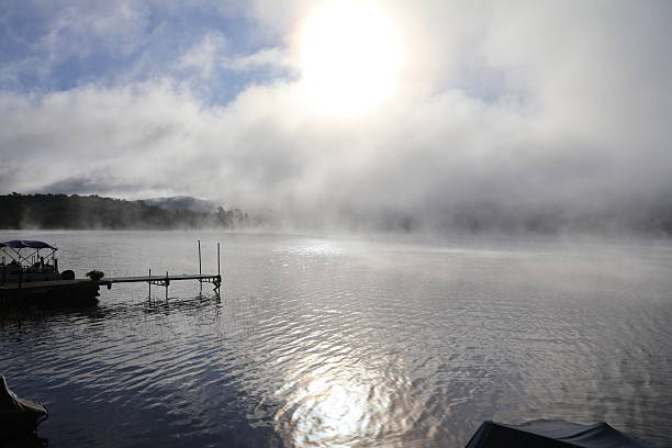 A dock front view of a placid looking lake glowing in the morning light as the fog evaporates under the heat of the sun.