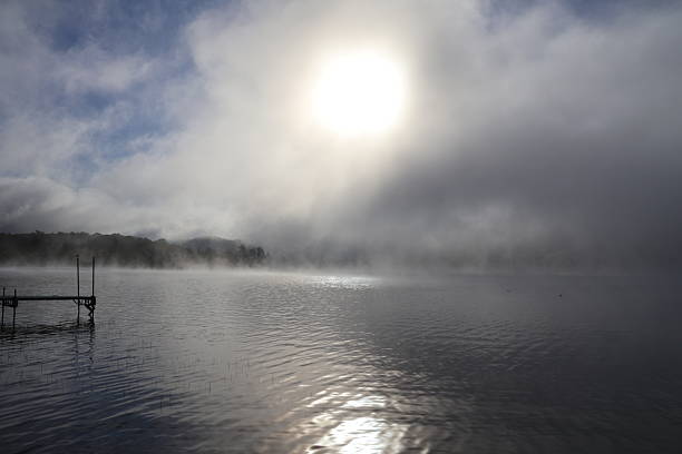 A dock front view of a placid looking lake glowing in the morning light as the fog evaporates under the heat of the sun.