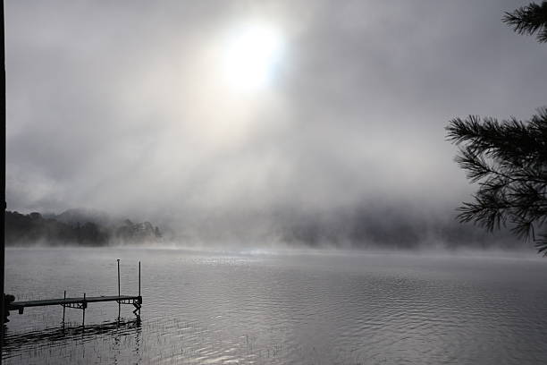 A dock front view of a placid looking lake glowing in the morning light as the fog evaporates under the heat of the sun.