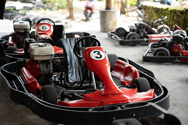 This picture shows a red go-kart parked indoors, with other go-karts visible in the background. The go-kart has a black seat and a white circular decal with the number \