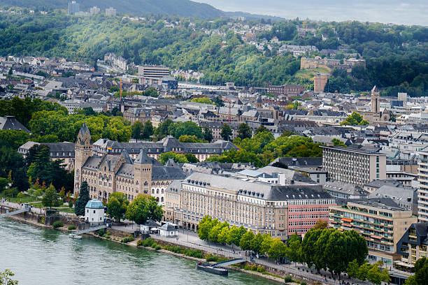 Koblenz Cityscape River Rhine Historic Architecture. Aerial view of Koblenz, Germany, with traditional European buildings, historic structures, and the green hills of Rhineland in the background. Captured on a cloudy summer day from above the riverbank, the image shows urban density, iconic architecture, and cultural heritage