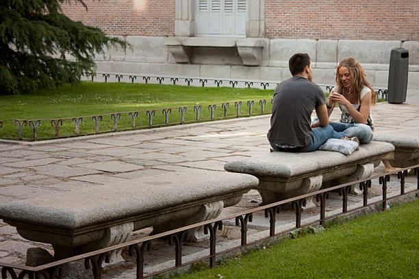 Madrid, Spain - May 13, 2011: Young man and woman sitting on a stone bench, looking at each other and talking, enjoying a casual moment together in a beautiful public park in Madrid, Spain