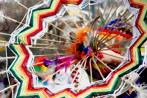 Hand holding an intricate handmade object, featuring vibrant rainbow yarn and natural feathers, creating a folk art craft or dreamcatcher at a market in Madrid