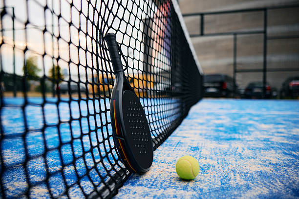Padel racket leaning against a black net on a vivid blue court with a yellow ball on the ground, suggesting a pause after play or the end of a match, close-up perspective