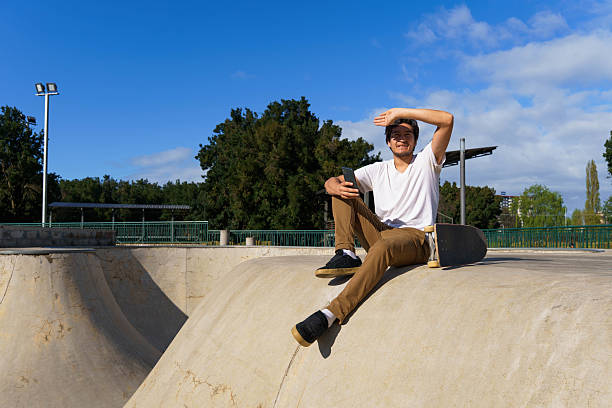 Young man sitting in a skatepark, shielding eyes from the sun and holding a smartphone