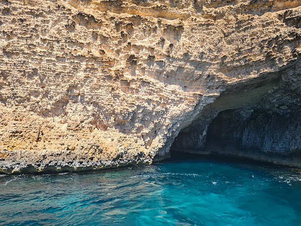 A rugged limestone cliff with a dark sea cave opens onto clear blue Mediterranean waters in Malta. The sunlight accentuates the unique textures of the rock face, creating a natural contrast with the deep shadows of the cave