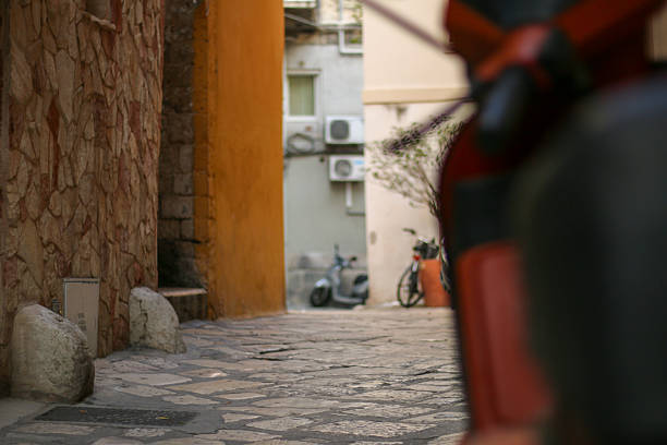 A narrow stone-paved alley in Bari, Italy, leads to a small courtyard with parked scooters and bicycles. Warmly colored walls and urban details create an authentic southern Italian atmosphere, evoking daily life in the historic city center.
