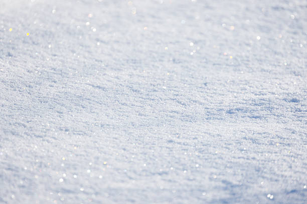 A sunlit, snow-covered ground glistens with tiny crystals, creating a tranquil winter scene. Natural background photo texture of a snowdrift with selective soft focus