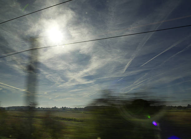 A scenic view of the English countryside captured from a moving train, showing a bright sun filtered through high clouds and contrails in a blue sky. Power lines and motion blur from trees emphasize the sense of travel and movement. Ideal for themes of transportation, commuting, rural landscapes, or scenic journeys.