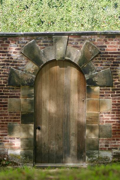 istock A rustic wooden arched door is set into a red brick wall with a stone surround. The setting is peaceful, with soft sunlight filtering through trees, creating a quiet, secluded atmosphere. 2238611693