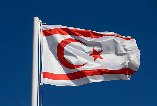 The national flag of Northern Cyprus on a pole, waving in the wind against a cloudy blue sky.