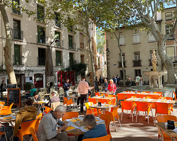 on a paved square in Ceret, a small town in the Pyrénées-Orientales. September 2025People sitting at tables and eating outside bars and restaurants in the shadows of plane trees in Place des Neuf Jet a public square in the town of Céret, France. The square is paved with stone and features a number of outdoor tables and chairs from nearby restaurants and cafes. September 2025