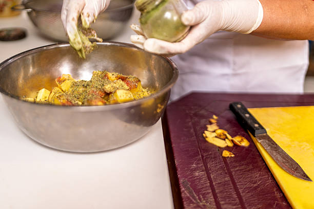 Chef hands wearing gloves seasoning chopped potatoes with various herbs and spices in a stainless steel bowl for cooking