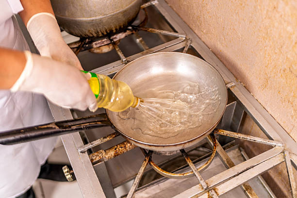 Chef in hygienic gloves pouring cooking oil from a plastic bottle into a metal frying pan on a gas stove in a restaurant kitchen