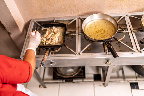 Chef stirring food in a griddle pan while another pan with oil heats on a commercial stove in a small kitchen