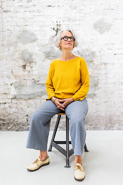 Senior woman sitting at an outdoor cafe table, looking thoughtful. A slice of cake and a cup of flowers are on the table, with a street scene in the background