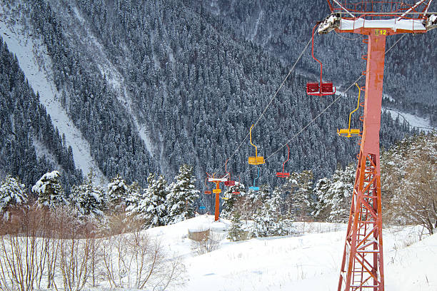 A colorful ski lift with bright seats passes through a snowy forest at the foot of the mountains. In the distance, tall snow-covered slopes covered with dense coniferous trees are visible